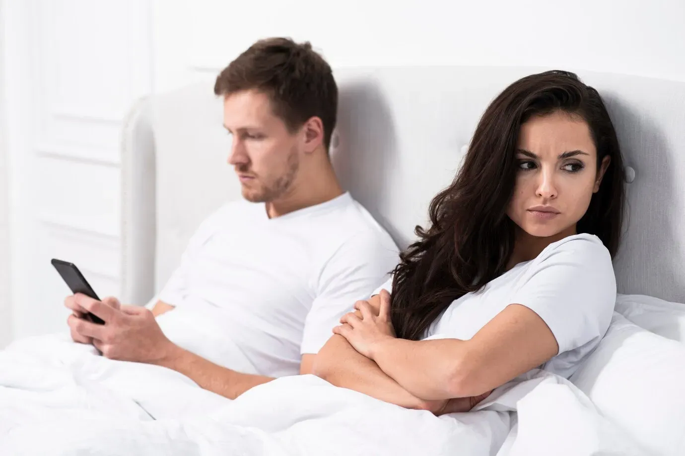 Woman in bed next to her husband on his phone